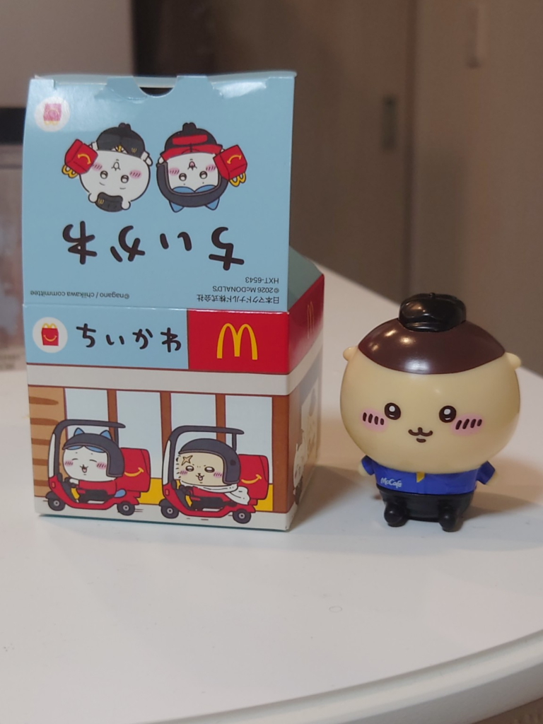 マクドナルドのちいかわハッピーセットの箱と、制服姿のキャラクターフィギュアを白い台の上に並べた写真。箱にはちいかわキャラクターとマクドナルドのデザインが描かれている。