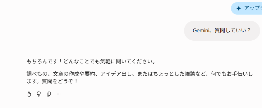 Gemini(AI)との日本語チャット画面のスクリーンショット。右上には部分的に青いUI要素「✦ アップ」が見える。右上のグレーの吹き出しにはユーザーのメッセージ「Gemini、質問していい?」とあり、その下のAIの返信には「もちろんです!どんなことでも気軽に聞いてください。調べもの、文章の作成や要約、アイデア出し、またはちょっとした雑談など、何でもお手伝いします。質問をどうぞ!」というテキストが続く。AIの返信の下には、いいね、よくないね、共有、その他のアクションアイコンが並んでいる。