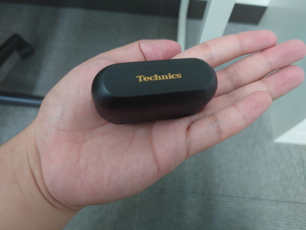 手のひらに黒いTechnicsのワイヤレスイヤホン充電ケースを乗せている写真。ケース上部には金色のTechnicsロゴがあり、背景には室内の床や家具がぼんやり写っている。